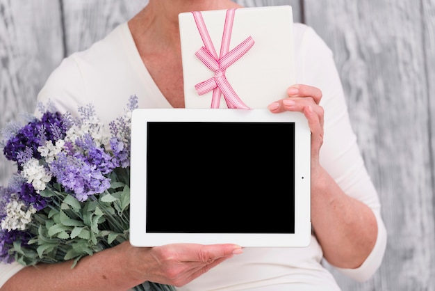 Section médiane d&#39;une femme tenant une boîte-cadeau; bouquet de fleurs et tablette numérique écran blanc à la main