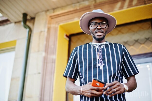 Séduisant homme afro-américain en chapeau de chemise rayée et lunettes de soleil marchant dans la rue au centre-ville