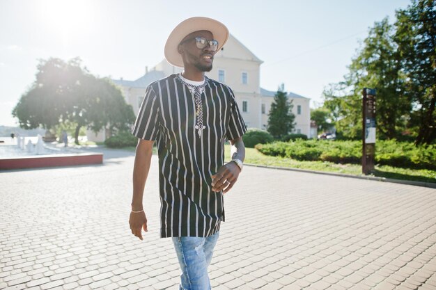 Séduisant homme afro-américain en chapeau de chemise rayée et lunettes de soleil marchant dans la rue au centre-ville