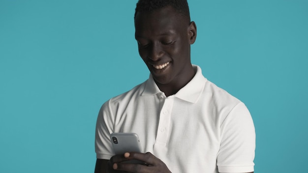 Séduisant homme afro-américain souriant textos avec des amis sur smartphone isolé sur fond bleu. Concept de technologie moderne