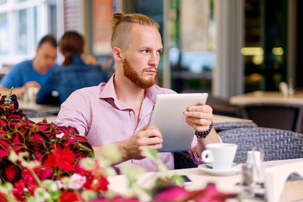 Un séduisant homme rousse barbu vêtu d'une chemise rose est assis à la table d'un café et utilise une tablette PC.