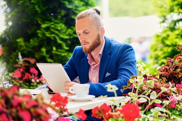 Un séduisant homme rousse barbu vêtu d'une chemise rose et d'une veste bleue est assis à la table d'un café et utilise une tablette PC.