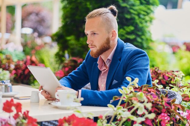 Un séduisant homme rousse barbu vêtu d'une chemise rose et d'une veste bleue est assis à la table d'un café et utilise une tablette PC.