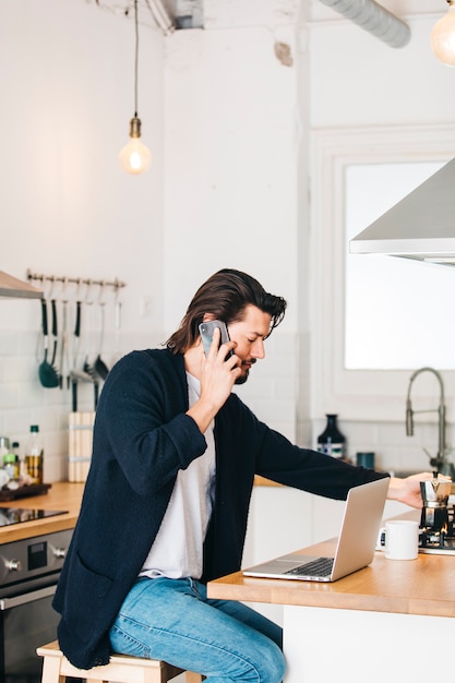 Un séduisant jeune homme assis dans la cuisine, parler au téléphone cellulaire