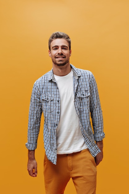 Séduisant jeune homme barbu en chemise à carreaux bleu et t-shirt blanc a l'air sûr de lui et pose sur fond orange