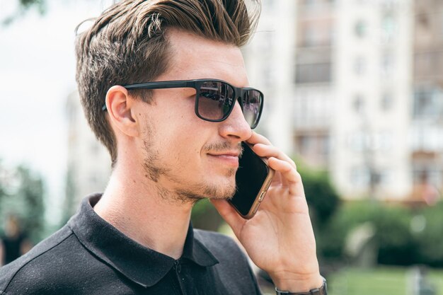 Séduisant jeune homme à lunettes de soleil parlant sur un smartphone dans la ville