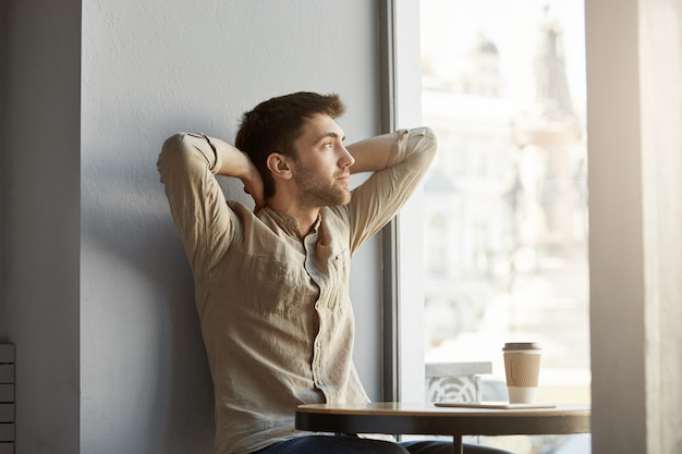 Photo gratuite séduisant jeune homme mal rasé assis dans un café, boire du café, regardant la fenêtre avec les mains derrière la tête, épuisé après une réunion d'affaires