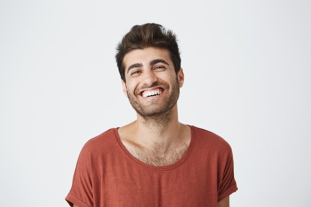 Séduisant jeune homme à la peau sombre non rasé en t-shirt rouge largement souriant en riant d'une image drôle sur Internet. Expressions faciales et émotions positives