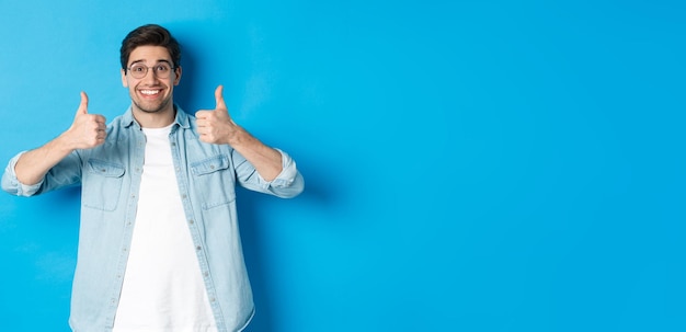 Séduisant jeune homme portant des lunettes et des vêtements décontractés montrant les pouces vers le haut en signe d'approbation comme quelque chose