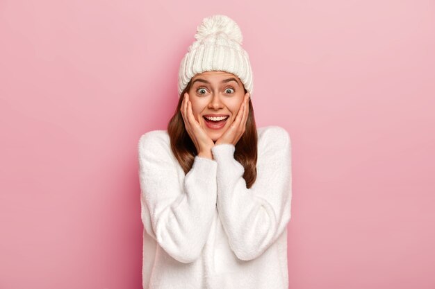 Séduisante belle femme brune se sent divertie et optimiste, touche doucement les joues, a un regard enthousiaste, porte un chapeau blanc chaud et un pull surdimensionné