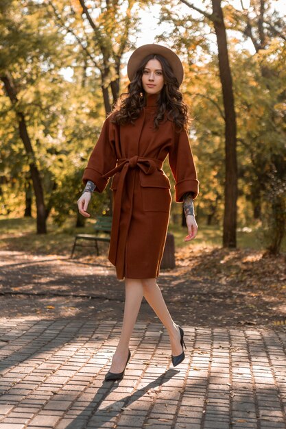 Séduisante élégante femme maigre souriante aux cheveux bouclés marchant dans le parc habillé en manteau brun chaud, style de rue à la mode automne