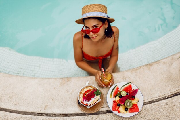 Séduisante Femme Brune Bronzée En Lunettes De Soleil Yeux De Chat Rouge Et Chapeau De Paille Relaxant Dans La Piscine Avec Assiette De Fruits Exotiques Pendant Les Vacances Tropicales. Tatouage élégant.