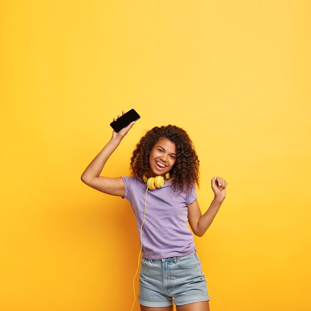 Séduisante femme positive détendue avec une coiffure afro, écoute de la musique dans des écouteurs, chante une chanson, lève les bras, bénéficie d'une qualité sonore impressionnante