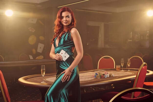 Une séduisante fille rousse vêtue d'une longue robe en satin bleu sourit et pose sur le côté avec deux as à la main contre une table de poker dans un casino de luxe. Passion, cartes, jetons, alcool, victoire, jeu - c'est moi