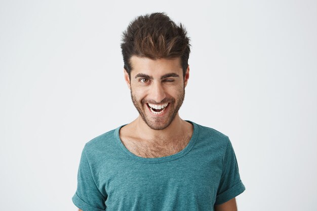 Séduisante et gaie jeune homme avec barbe et coupe de cheveux élégante portant un t-shirt bleu clignant des yeux avec plaisir ayant une expression heureuse. Expressions faciales et concept d'émotions de personnes