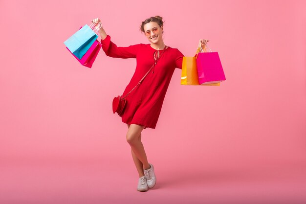 Séduisante heureuse souriante femme élégante accro du shopping en robe rouge à la mode tenant des sacs colorés sur un mur rose isolé, vente excité, tendance de la mode printemps été