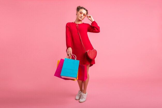 Séduisante heureuse souriante femme élégante accro du shopping en robe rouge à la mode tenant des sacs colorés sur un mur rose isolé, vente excité, tendance de la mode printemps été