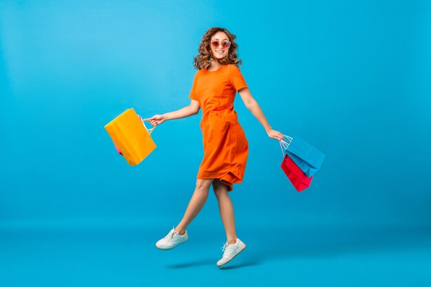 Séduisante heureuse souriante femme élégante accro du shopping en robe surdimensionnée à la mode orange sautant tenant des sacs à provisions sur fond bleu studio isolé