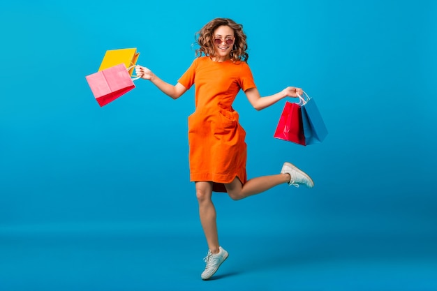 Séduisante heureuse souriante femme élégante accro du shopping en robe surdimensionnée à la mode orange sautant tenant des sacs à provisions sur fond bleu studio isolé