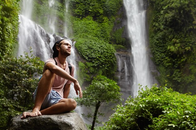 Séduisante jeune aventurier barbu ne portant pas de chaussures ayant une pause sur un gros rocher lors d'une randonnée seule dans la forêt tropicale. Randonneur élégant se détendre à l'extérieur dans la jungle avec une cascade incroyable