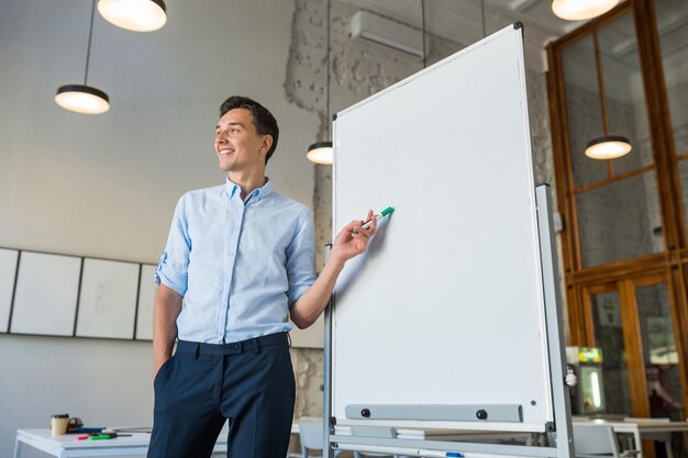 Séduisante jeune bel homme souriant debout au tableau blanc vide avec marqueur