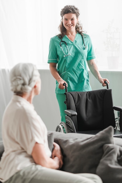 Séduisante jeune femme infirmière apportant un fauteuil roulant à un patient senior