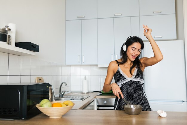 Séduisante jeune femme souriante maigre s'amusant à cuire des œufs à la cuisine le matin en prenant le petit déjeuner habillé en pyjama, écoutant de la musique sur des écouteurs dansant