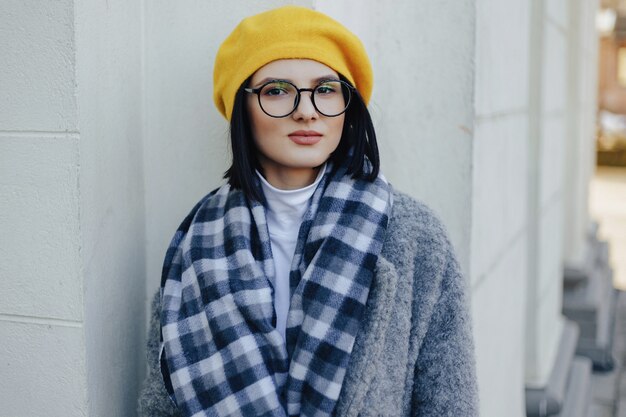 Séduisante jeune fille à lunettes en manteau et béret jaune sur une surface légère simple