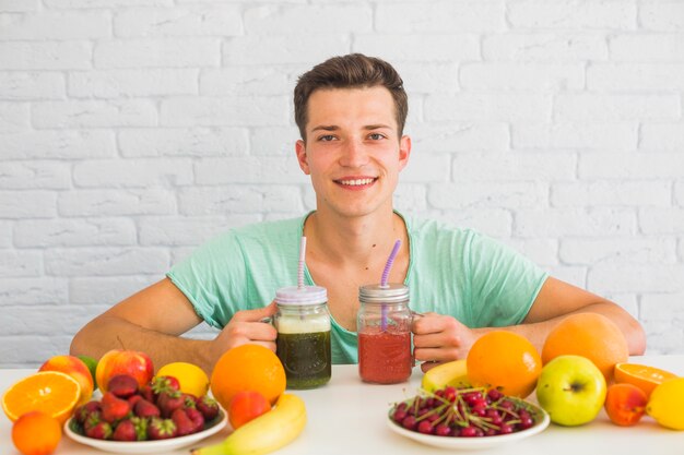 Séduisante jeune homme tenant des pots de smoothie vert et rouge avec des fruits colorés sur la table