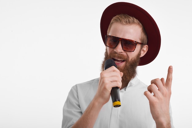 Séduisante jeune showman barbu portant des lunettes de soleil élégantes et un chapeau tenant le microphone et levant le doigt avant tout en annonçant la performance du chanteur populaire