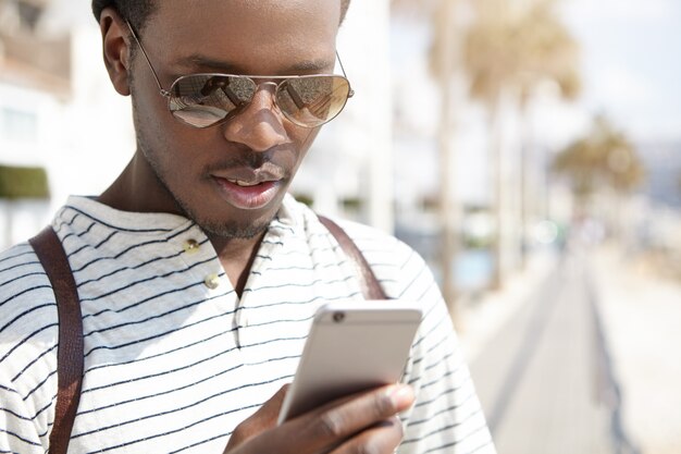 Séduisante jeune voyageur afro-américain dans des tons à la mode utilisant l'application de navigation sur son téléphone mobile générique, à la recherche d'une direction tout en marchant seul dans une ville étrangère. Les gens et la technologie moderne