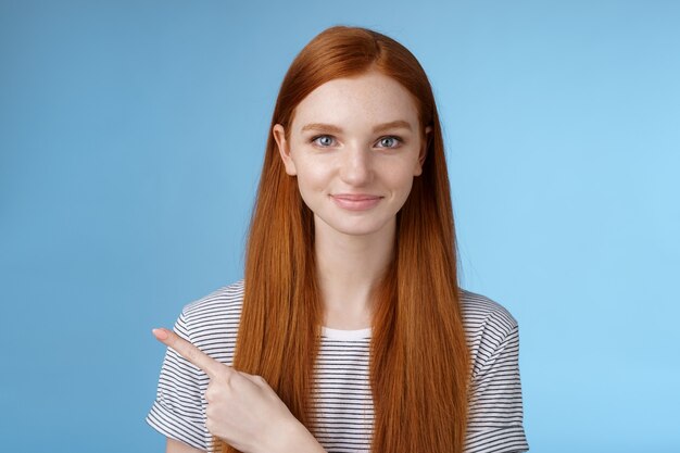 Séduisante petite amie rousse belle et modeste agissant mature amicale souriante pointant l'index gauche donnant la direction montrer comment salle de bain debout fond bleu joyeux sourire aimable