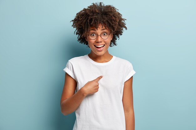 Sélectionnez-moi s'il vous plaît. Une femme joyeuse à la peau sombre se pointe du doigt, ne peut pas croire sa chance, porte un t-shirt décontracté, isolée sur un mur bleu, dit que je suis ce dont vous avez besoin.