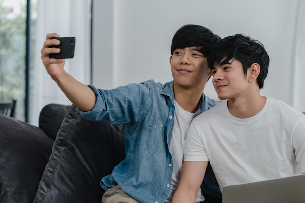 Selfie drôle jeune couple gay romantique par téléphone portable à la maison. Asiatique amant mâle heureux se détendre avec technologie téléphone mobile souriant prendre une photo ensemble en position couchée dans le salon.