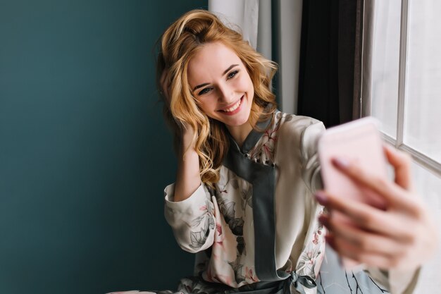 Selfie du matin de la belle jeune femme, fille heureuse et joyeuse. Elle porte un pyjama en soie, a les cheveux blonds ondulés, assise sur le rebord de la fenêtre dans une chambre avec un mur turquoise.