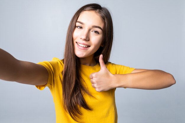 Selfie de femme heureuse montrant le pouce vers le haut et un clin de œil isolé sur mur blanc