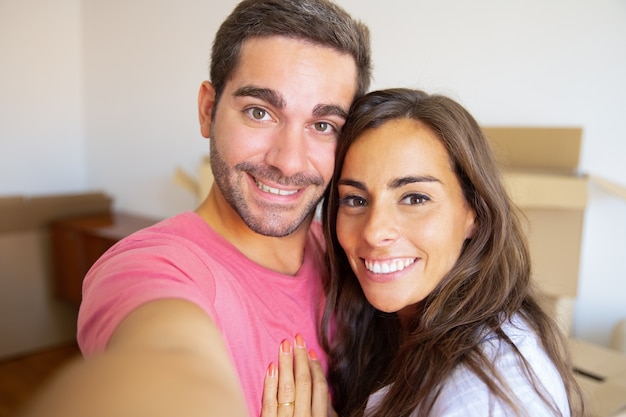 Selfie de l'heureux jeune couple dans leur nouvelle maison, posant avec des boîtes en carton en arrière-plan, tenant le gadget en main
