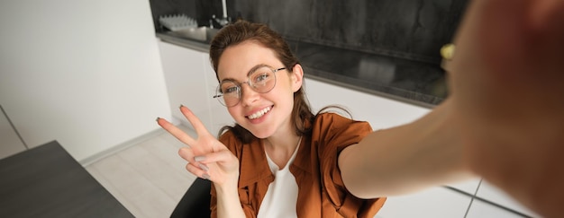 Photo gratuite selfie d'une jeune femme mignonne en lunettes posant à la maison dans la cuisine en prenant des photos sur son téléphone portable