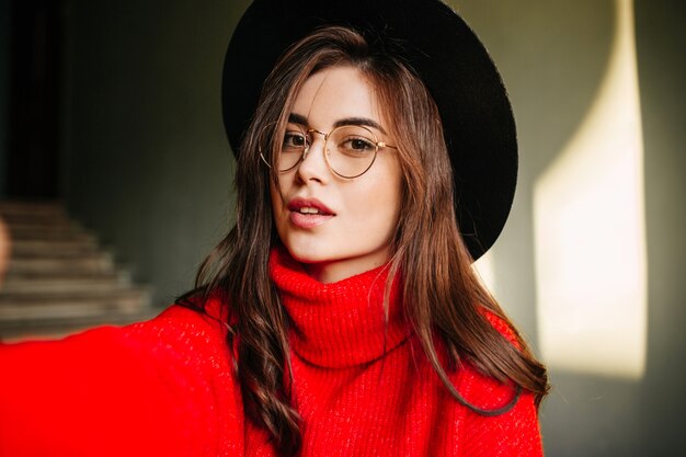 Selfie de jeune fille européenne aux cheveux noirs ondulés en pull rouge. Modèle en chapeau et lunettes posant