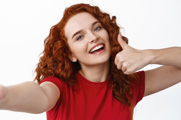 Selfie de jeune fille rousse heureuse avec une coiffure frisée montrant le pouce vers le haut, loue et recommande quelque chose de bien, souriant au smartphone, mur blanc