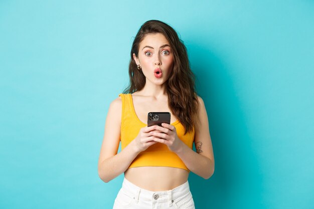 Semble intéressant. Jolie fille intriguée tenant un smartphone, regardant excité et disant wow après avoir lu une publicité en ligne, debout sur fond bleu