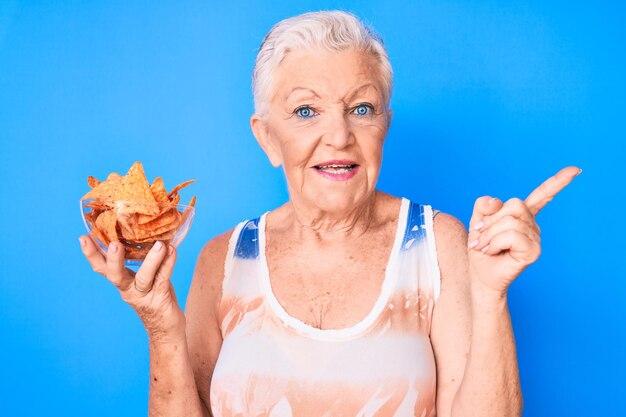 Photo gratuite senior belle femme aux yeux bleus et aux cheveux gris tenant des chips de pommes de terre nachos souriant heureux pointant avec la main et le doigt sur le côté