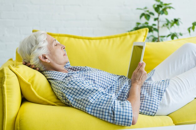 Senior femme allongée sur un canapé jaune en regardant une tablette numérique