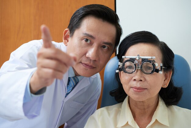 Senior femme asiatique portant une monture de lentille d'essai et ophtalmologiste pointant lors d'un bilan de la vue