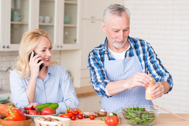 Senior femme souriante parlant au téléphone portable et son mari préparant la salade dans la cuisine