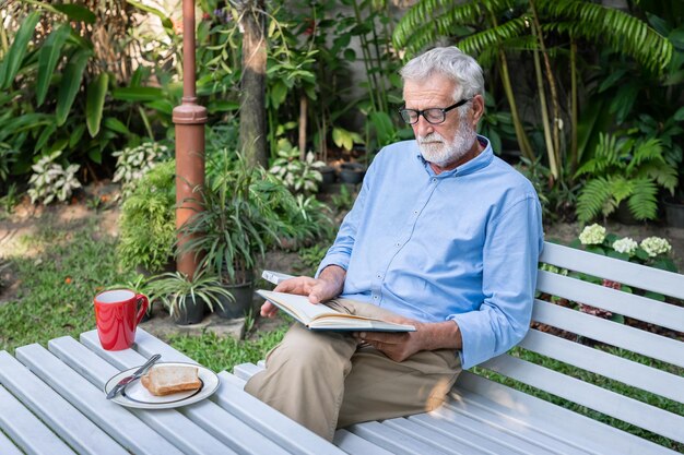 Senior homme âgé lisant un livre avec petit-déjeuner et tasse de café dans le jardin