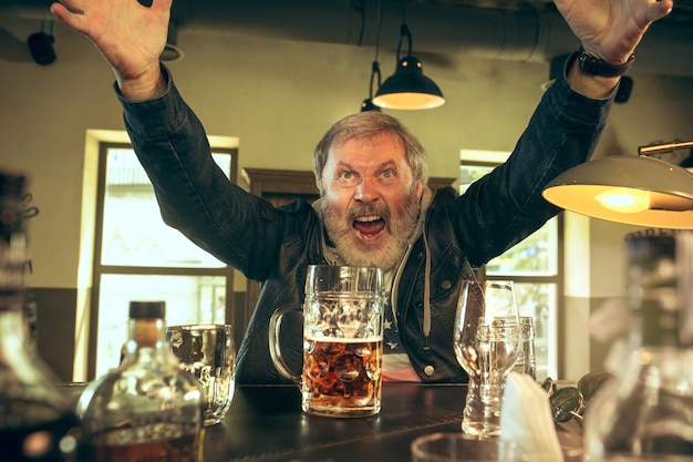 Photo gratuite senior homme barbu, boire de l'alcool dans un pub et regarder un programme sportif à la télévision. profitant de ma bière et de ma bière préférées. homme avec chope de bière assis à table. fan de football ou de sport. concept d'émotions humaines