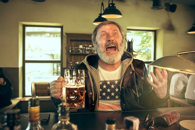 Photo gratuite senior homme barbu, boire de l'alcool dans un pub et regarder un programme sportif à la télévision. profitant de ma bière et de ma bière préférées. homme avec chope de bière assis à table. fan de football ou de sport. concept d'émotions humaines