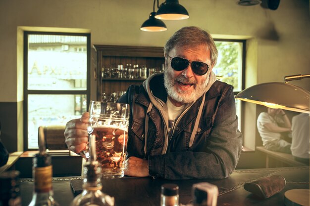 Senior homme barbu, boire de l'alcool dans un pub et regarder un programme sportif à la télévision. Profitant de ma bière et de ma bière préférées. Homme avec chope de bière assis à table. Fan de football ou de sport. Concept d'émotions humaines