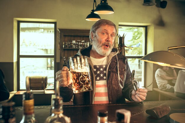 Senior homme barbu, boire de l'alcool dans un pub et regarder un programme sportif à la télévision. Profitant de ma bière et de ma bière préférées.